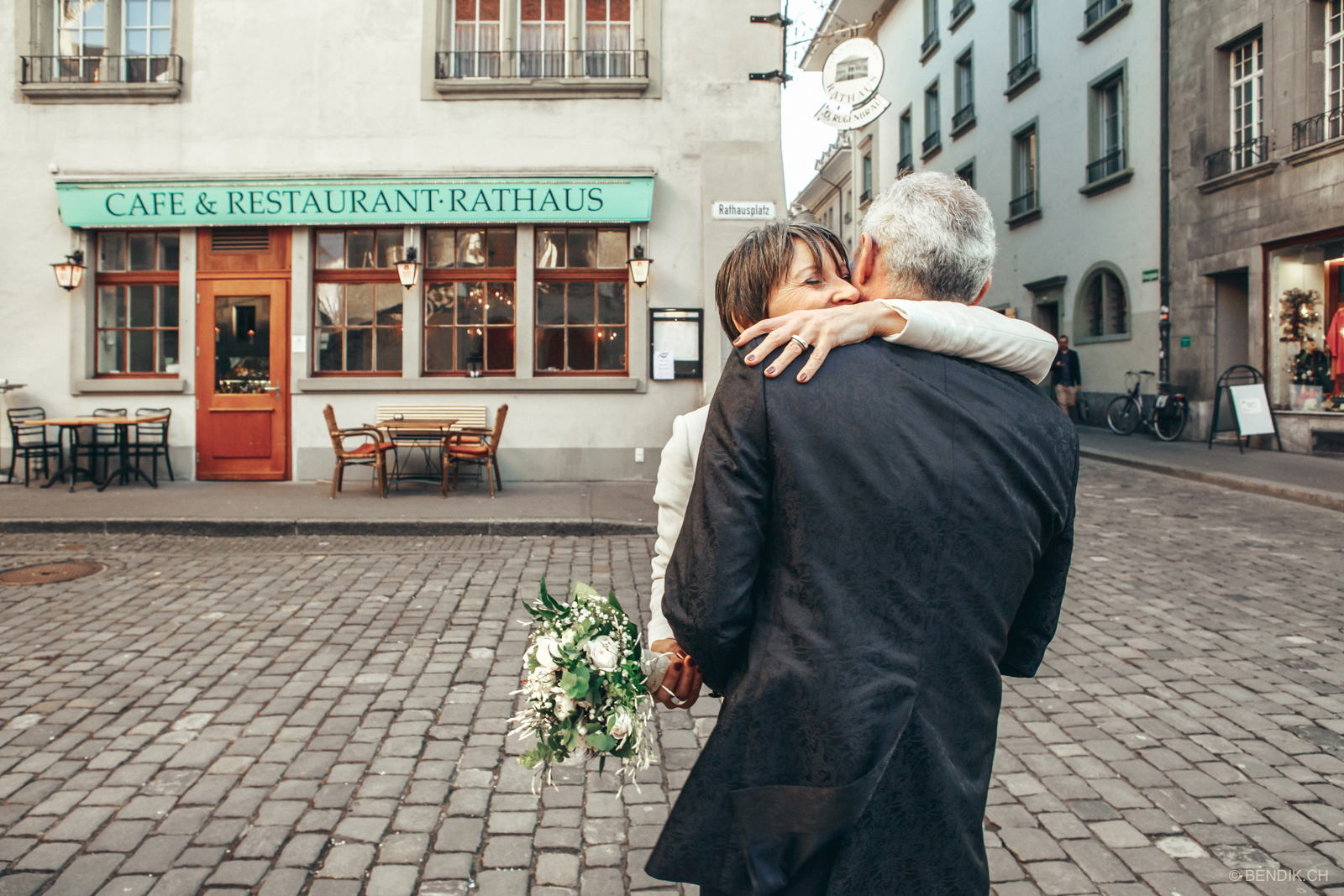 Brautpaar umarmt sich mit kleinem Kaffeehaus im Hintergrund